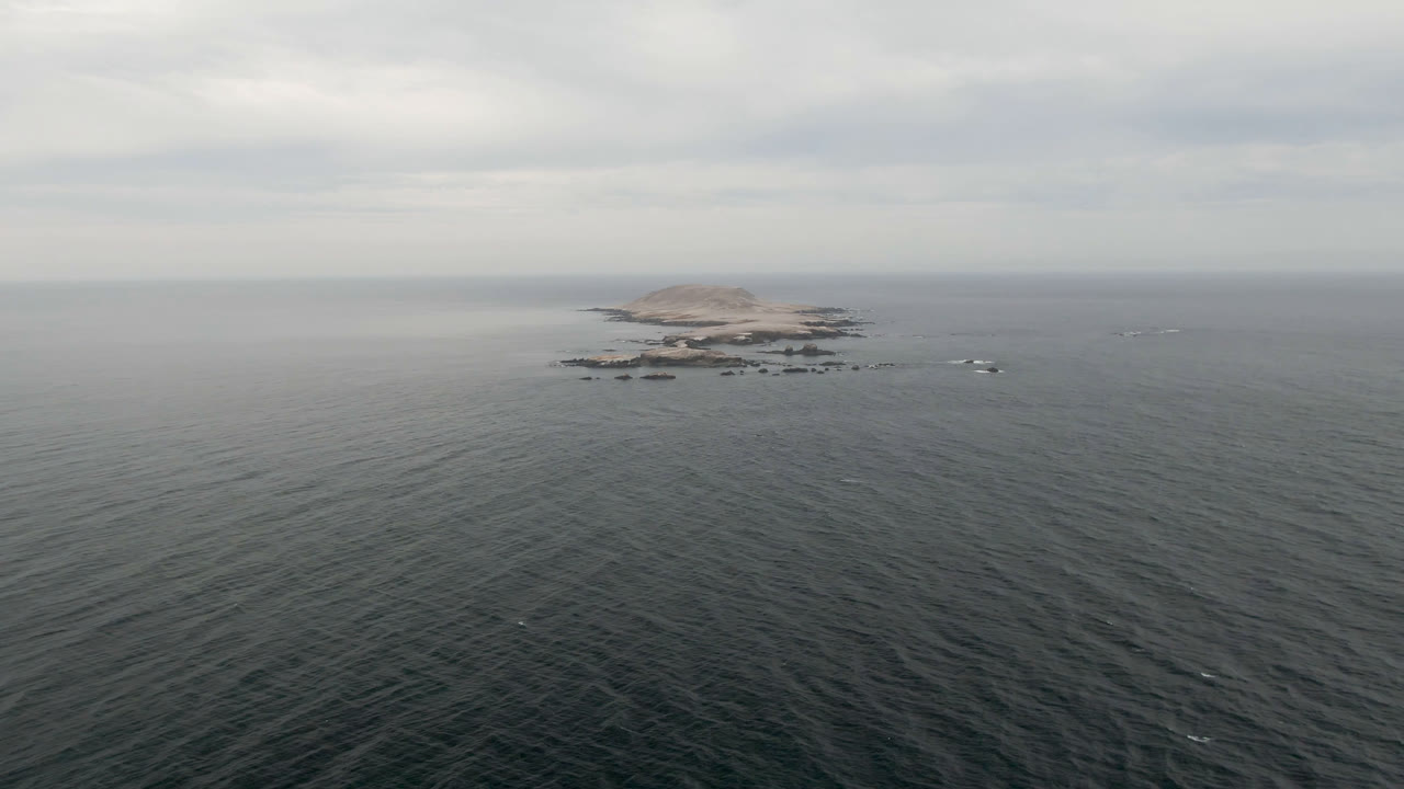 A distant rocky island sits near the horizon beyond dark rippled ocean water in Bahia Ascuncion, Mexico, under gray cloud cover with no visible people.