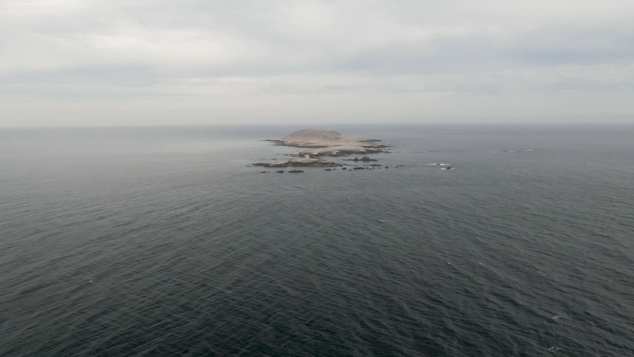 The centered island remains near the horizon in Bahia Ascuncion, Mexico, with dark textured ocean in the foreground and pale gray clouds above, no visible people.