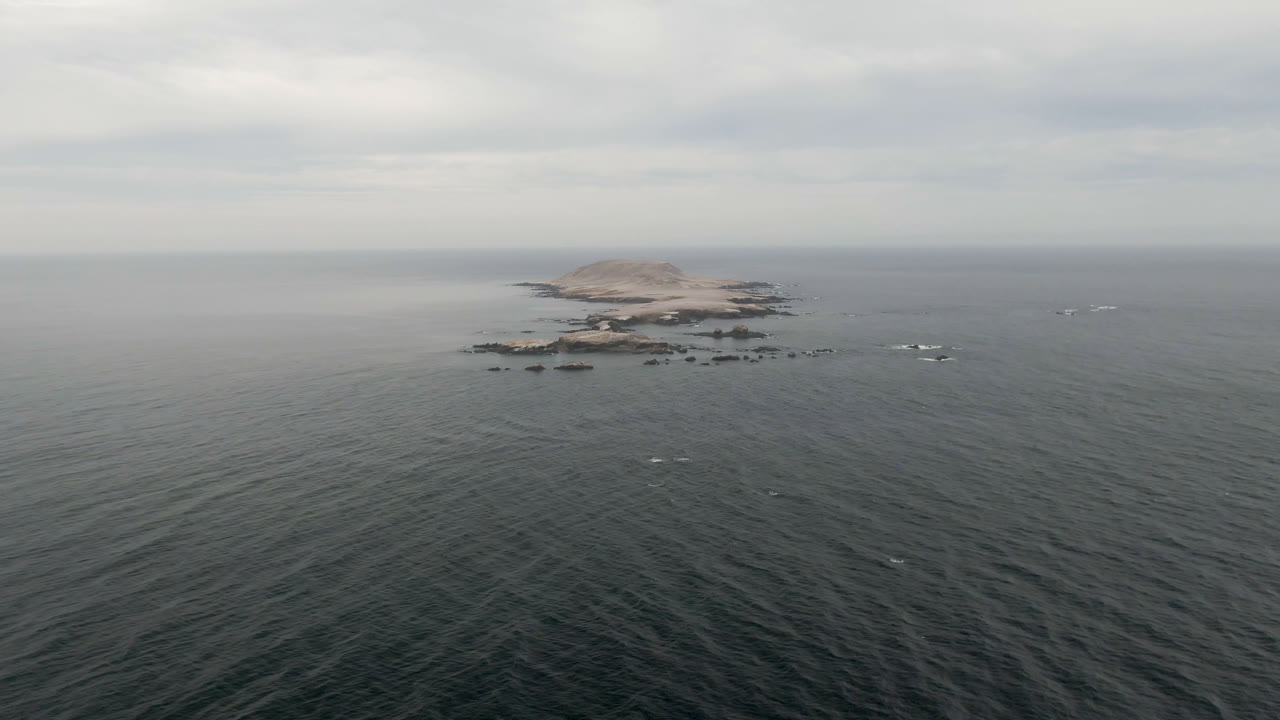 Wide aerial view in Bahia Ascuncion, Baja California Sur, Mexico, with dark ocean foreground, a small rocky island near the horizon, scattered white surf around rocks, and no visible people.