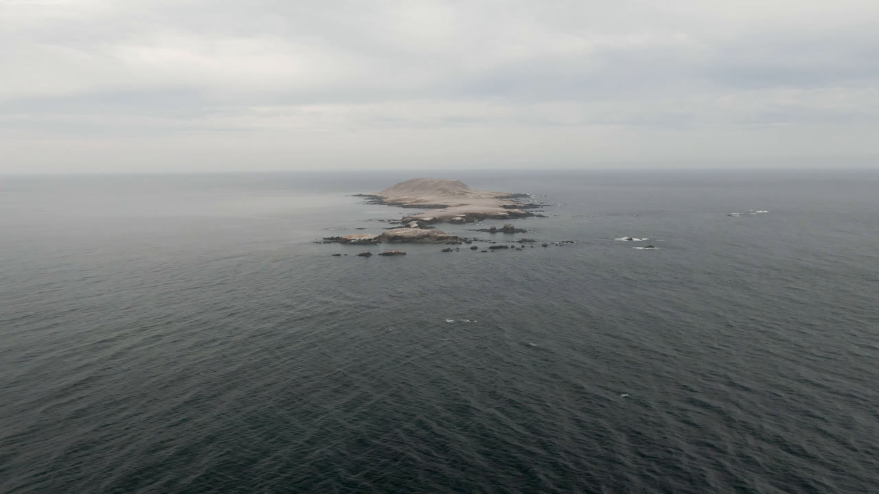 Aerial frame from Bahia Ascuncion showing textured green-gray water in the foreground, the rocky island slightly left of center near the horizon, pale cloud layers above, and no visible people.
