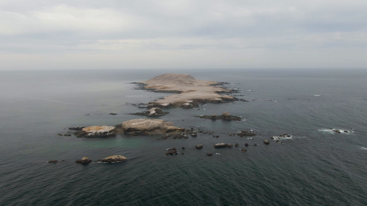 Aerial view over Bahia Ascuncion, Baja California Sur, with a pale rocky island near the horizon, scattered dark rocks in green-blue water, and no visible people.