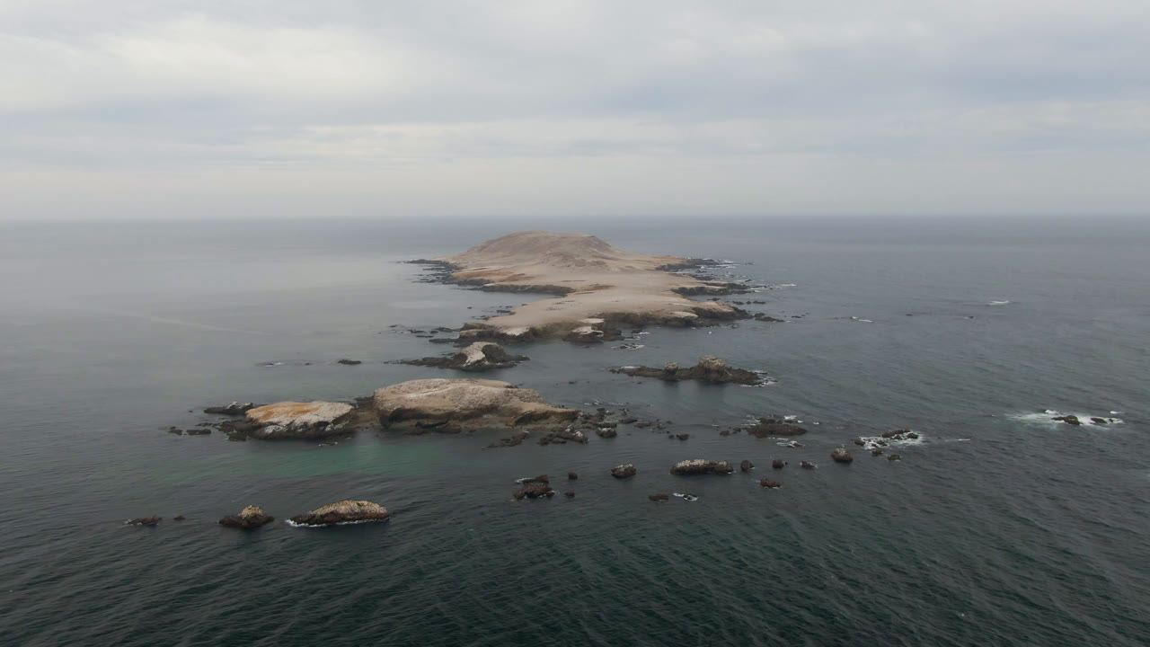 Aerial frame in Bahia Ascuncion showing the rocky island centered beyond a field of reef rocks, with muted daylight on the ocean and no visible people.