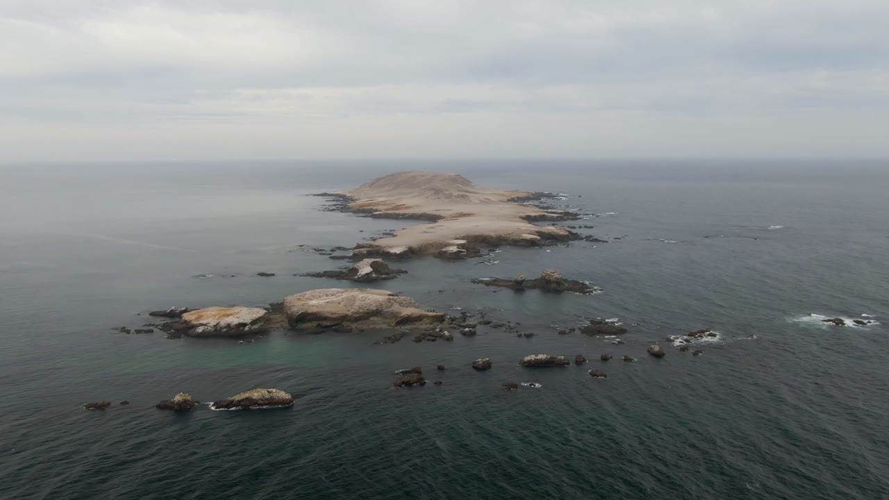Wide drone view of a rocky island and surrounding ocean under cloudy sky.