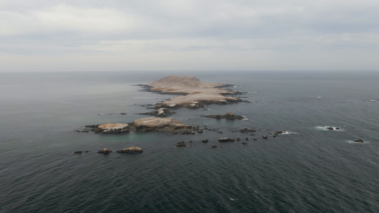 Wide aerial frame over Bahia Ascuncion, Baja California Sur, showing a low rocky island beyond scattered ocean rocks under gray cloud cover, with no visible people.