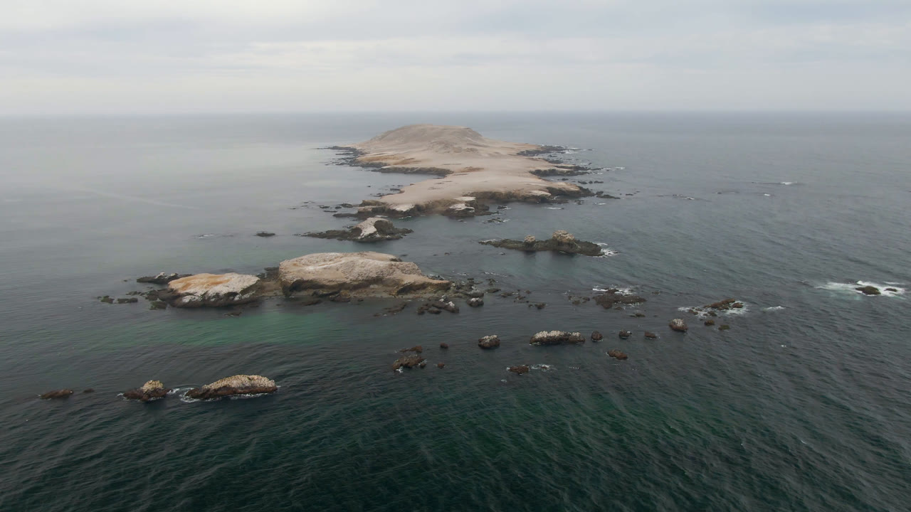 Aerial view near Bahia Ascuncion, Baja California Sur, with a pale rocky island beyond scattered rocks, dark teal ocean in the foreground, and no visible people.