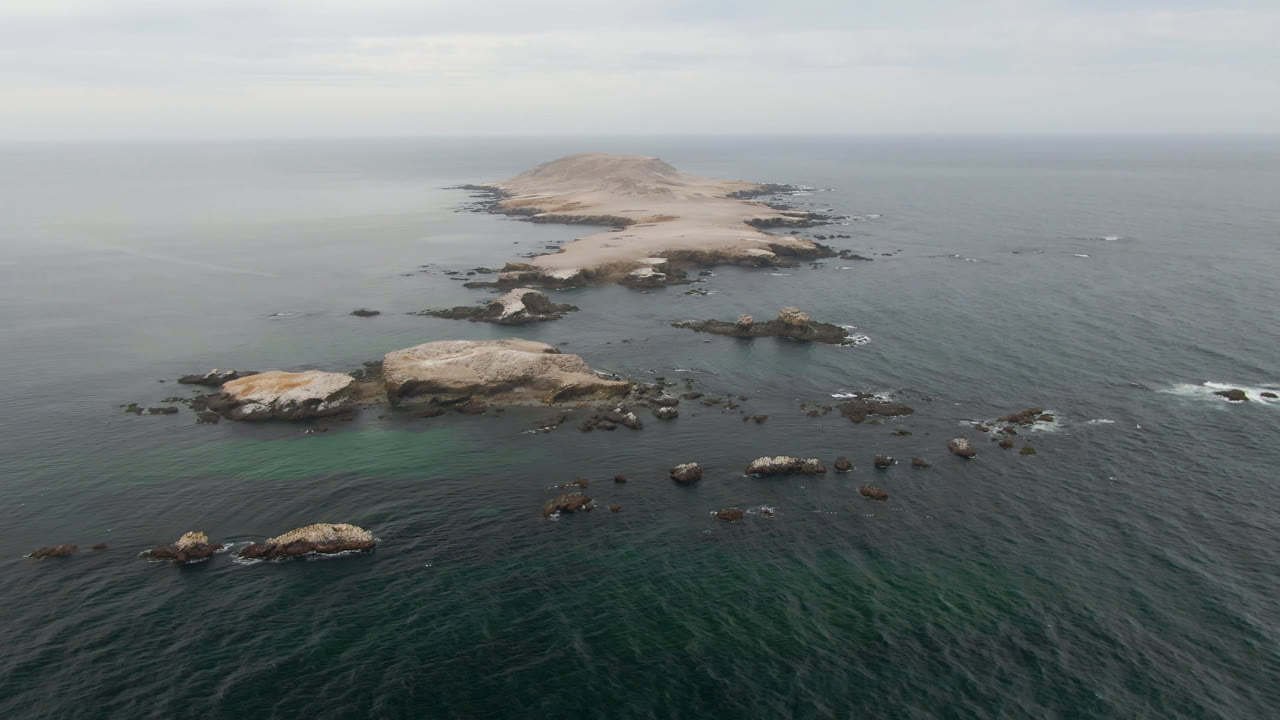 Wide aerial frame near Bahia Ascuncion showing the rocky island high in frame, reef-colored shallows around nearby rocks, open water below, and no visible people.