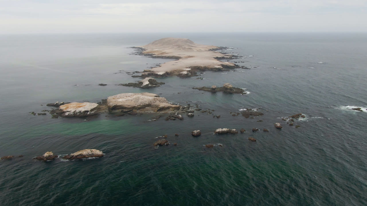 Drone-like aerial view over teal water toward a pale rocky island and low waves.