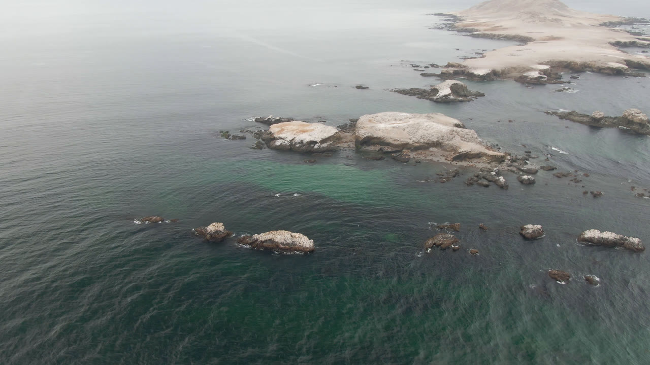 Opening aerial frame over Bahia Ascuncion with dark green water, pale submerged areas, scattered rocks, a low shoreline, a tiny light boat offshore, and no visible people.