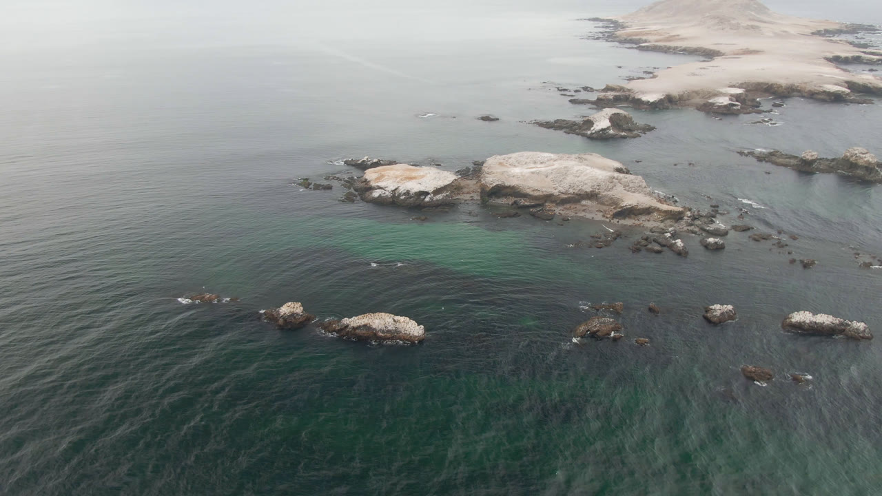 Aerial frame from Bahia Ascuncion showing calm green-gray water, rocky outcrops, a pale shallow patch near center, distant shoreline, a small light boat to the left, and no visible people.