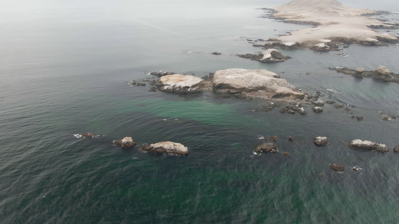 High aerial coastal view with dark water, pale underwater patch, rocks, and shoreline in haze.