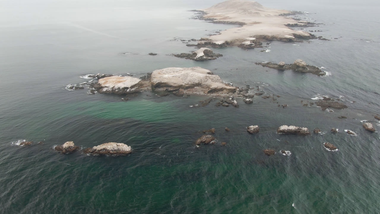 Aerial view in Bahia Ascuncion with darker water texture in the foreground, white foam around rocks, a sandy island high in frame, and no visible people.