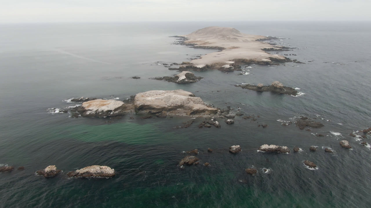 Opening aerial frame over Bahia Ascuncion water shows a low rocky island beyond scattered offshore rocks, with no visible people.