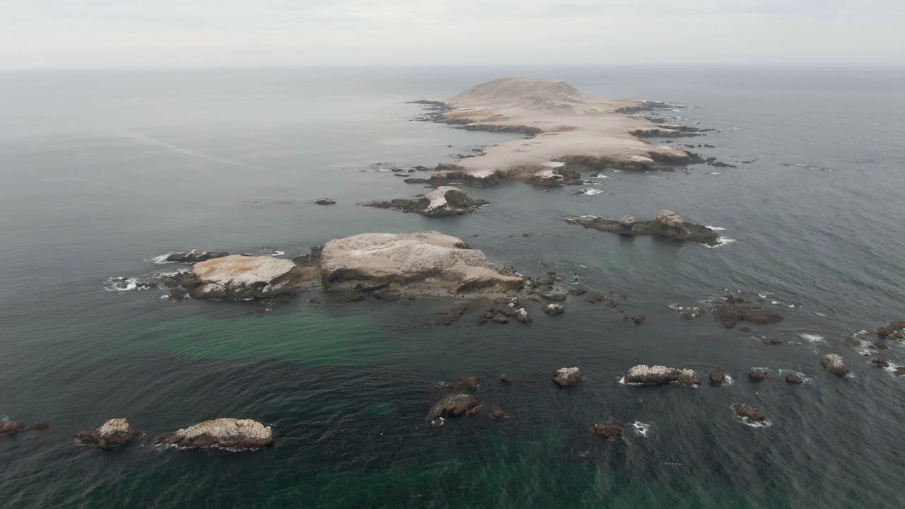 Aerial view in Bahia Ascuncion places pale foreground rocks below the rocky island, with muted ocean water and no visible people.