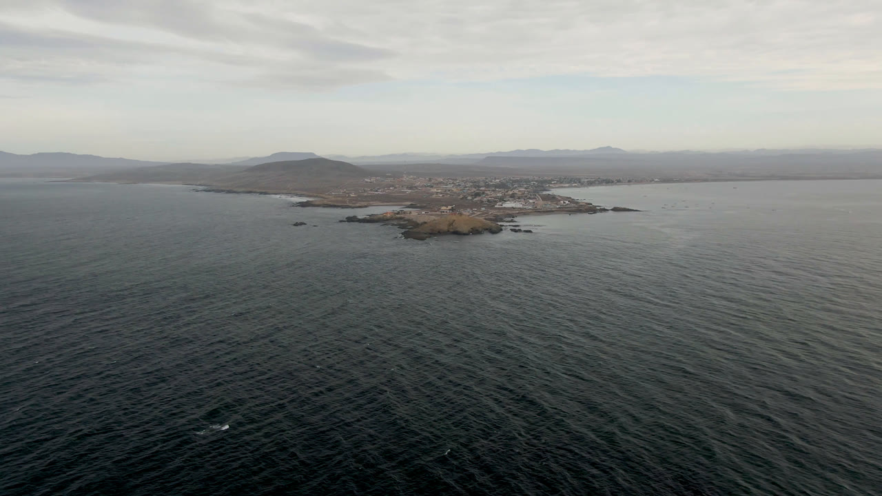 Opening aerial frame over dark Pacific water toward a small rocky island off Bahia Ascuncion, Baja California Sur, with distant hills under cloud cover and no visible people.