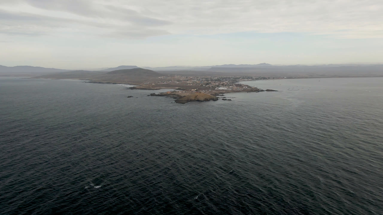 Aerial frame over rippled ocean with the small island slightly clearer in the midground, Bahia Ascuncion coastline beyond it, cloudy sky above, and no visible people.