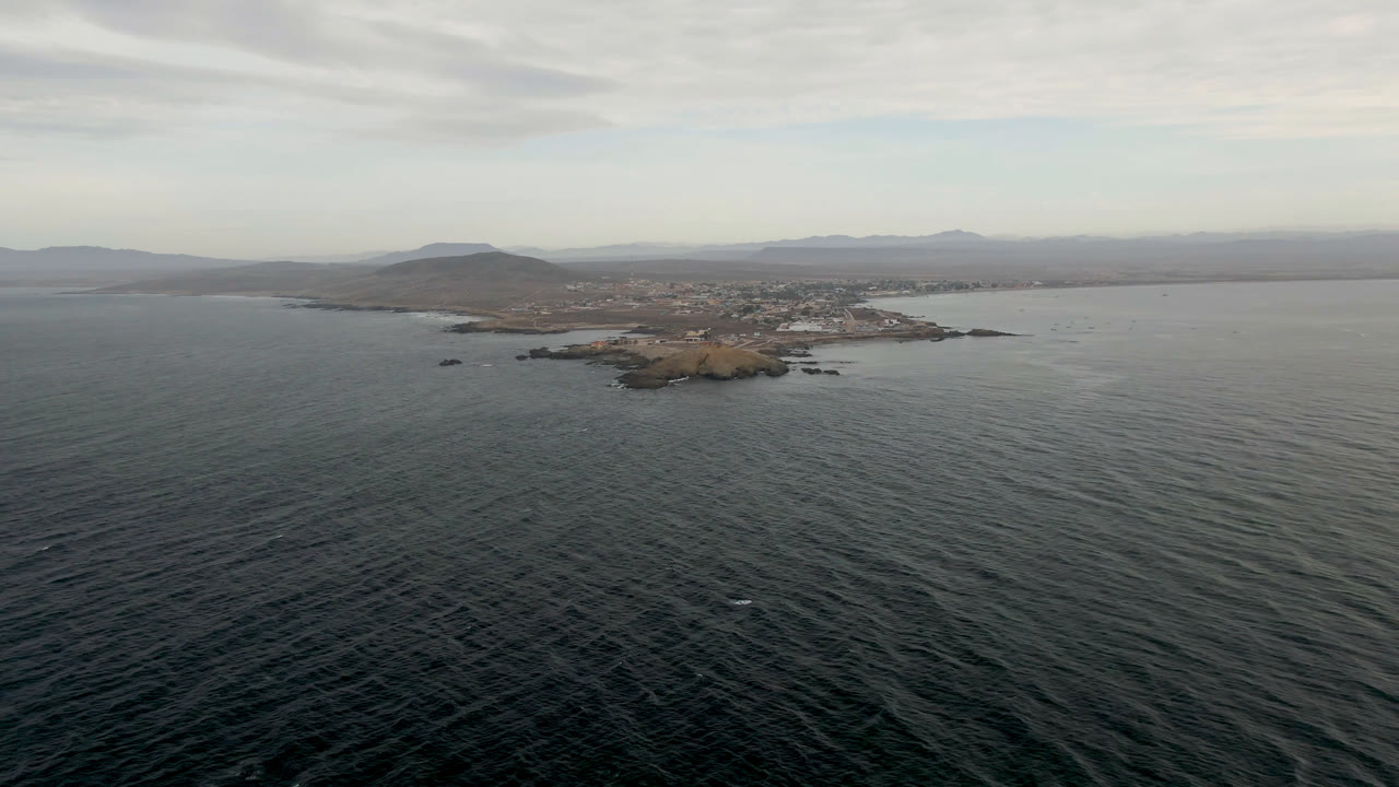 Aerial view over dark ocean toward a small rocky island and distant shoreline
