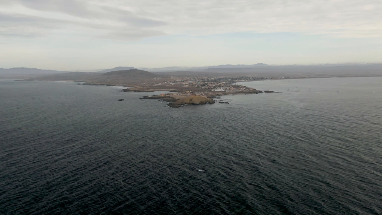 Drone-view frame from Bahia Ascuncion with textured ocean in the foreground, a rocky island left of center, a low coastal town beyond it, hazy hills, layered clouds, and no visible people.