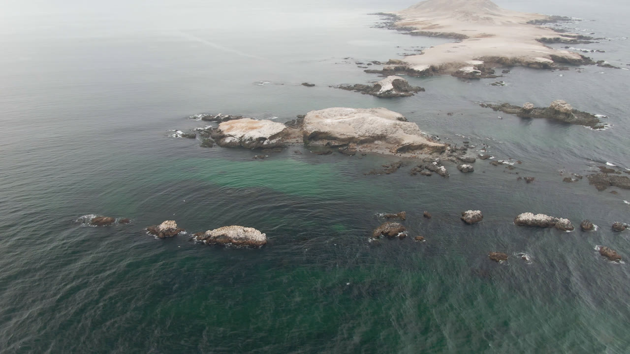 Aerial view over Bahia Ascuncion, Baja California Sur, with green-gray water, pale rock outcrops, scattered dark rocks, and no visible people at the opening frame.