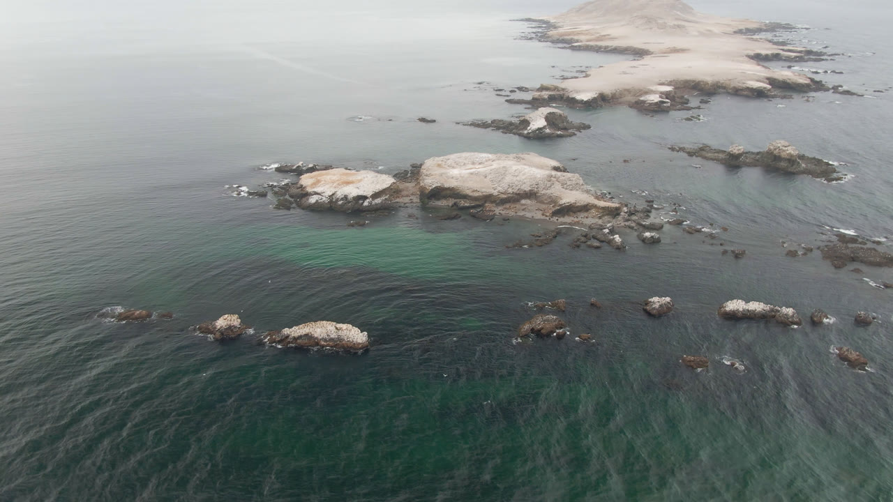 Aerial frame from Bahia Ascuncion showing pale midground rocks, darker stones around them, faint wave lines, and no visible people.