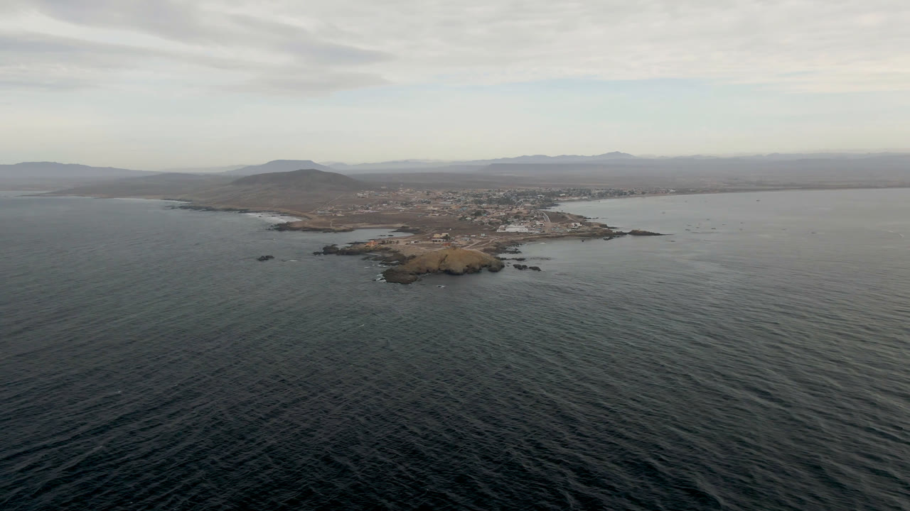 Wide aerial frame over dark rippled water toward a small rocky island off Bahia Ascuncion, Baja California Sur, with low town buildings and muted clouds in the background, no visible people.