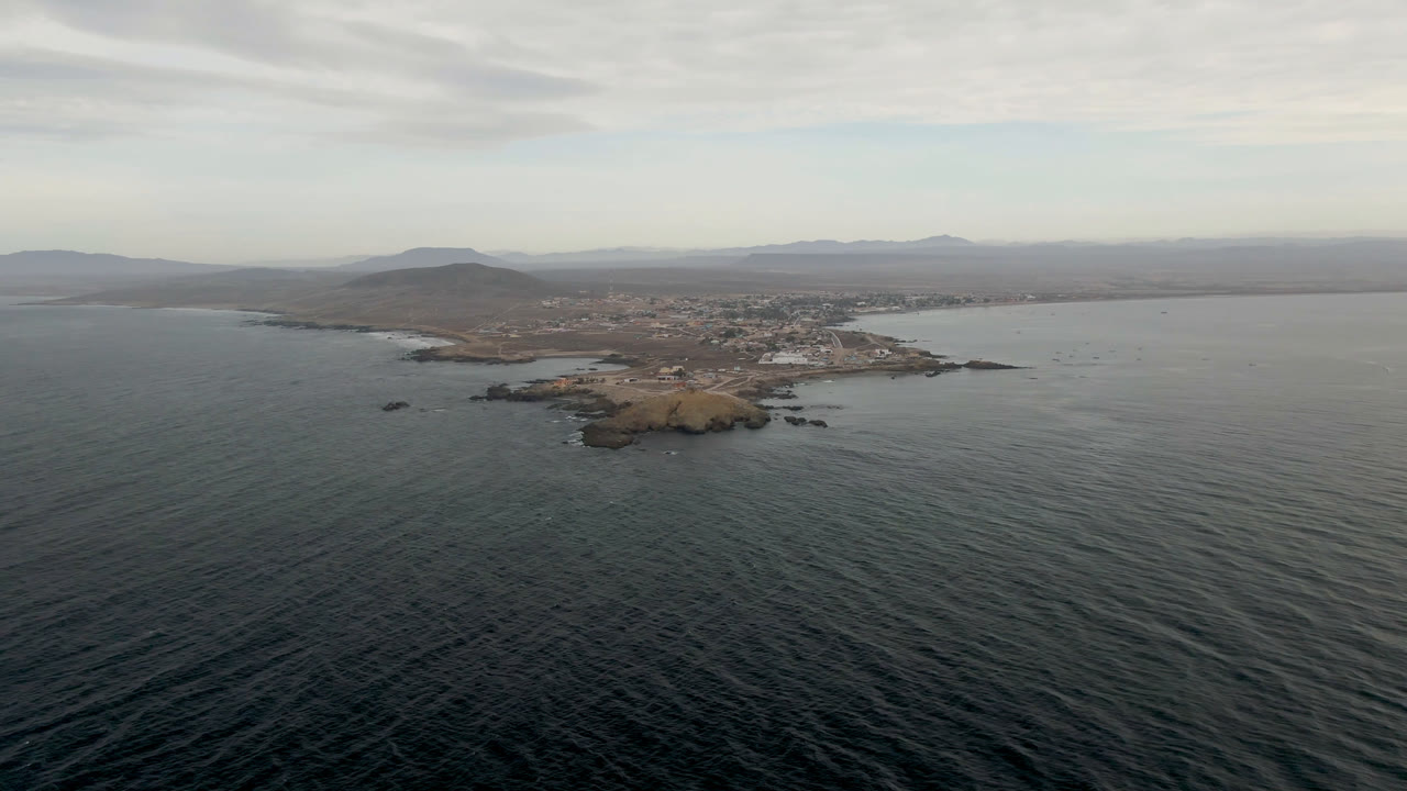Aerial view across textured ocean toward the offshore island near Bahia Ascuncion, with faint white surf on the rocks and a low gray cloud layer above the shore, no visible people.