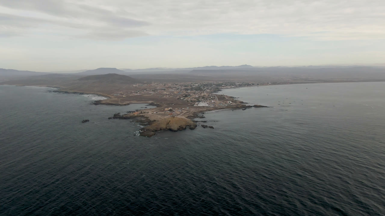 Wide aerial frame over Bahia Ascuncion, Baja California Sur, Mexico, with dark ocean in front, a tan rocky point near shore, a low town beyond, distant mountains, cloudy sky, and no visible people.