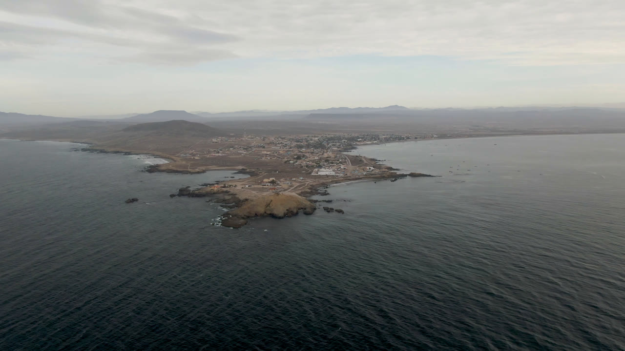 Aerial view over Bahia Ascuncion with textured dark water across the foreground, a rocky tan landform near the center, low buildings along the coast, hazy mountains, cloud cover, and no visible people.