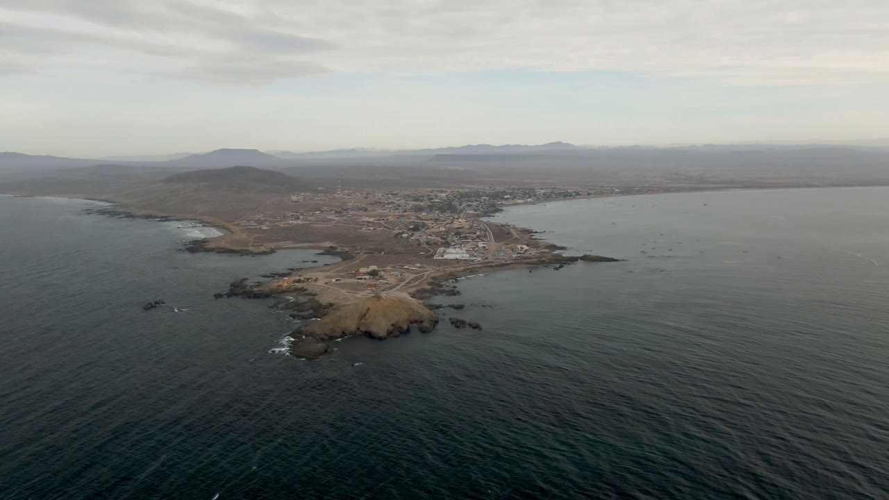 Aerial frame over Bahia Ascuncion, Mexico, with dark ocean in the foreground, a rocky point near center, the town along the bay, distant hills, and no visible people.
