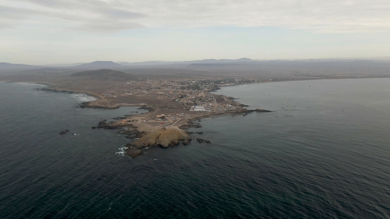 Wide aerial view of Bahia Ascuncion with the bay curving around a rocky shore, town buildings set back from the water, hazy inland hills, and no visible people.