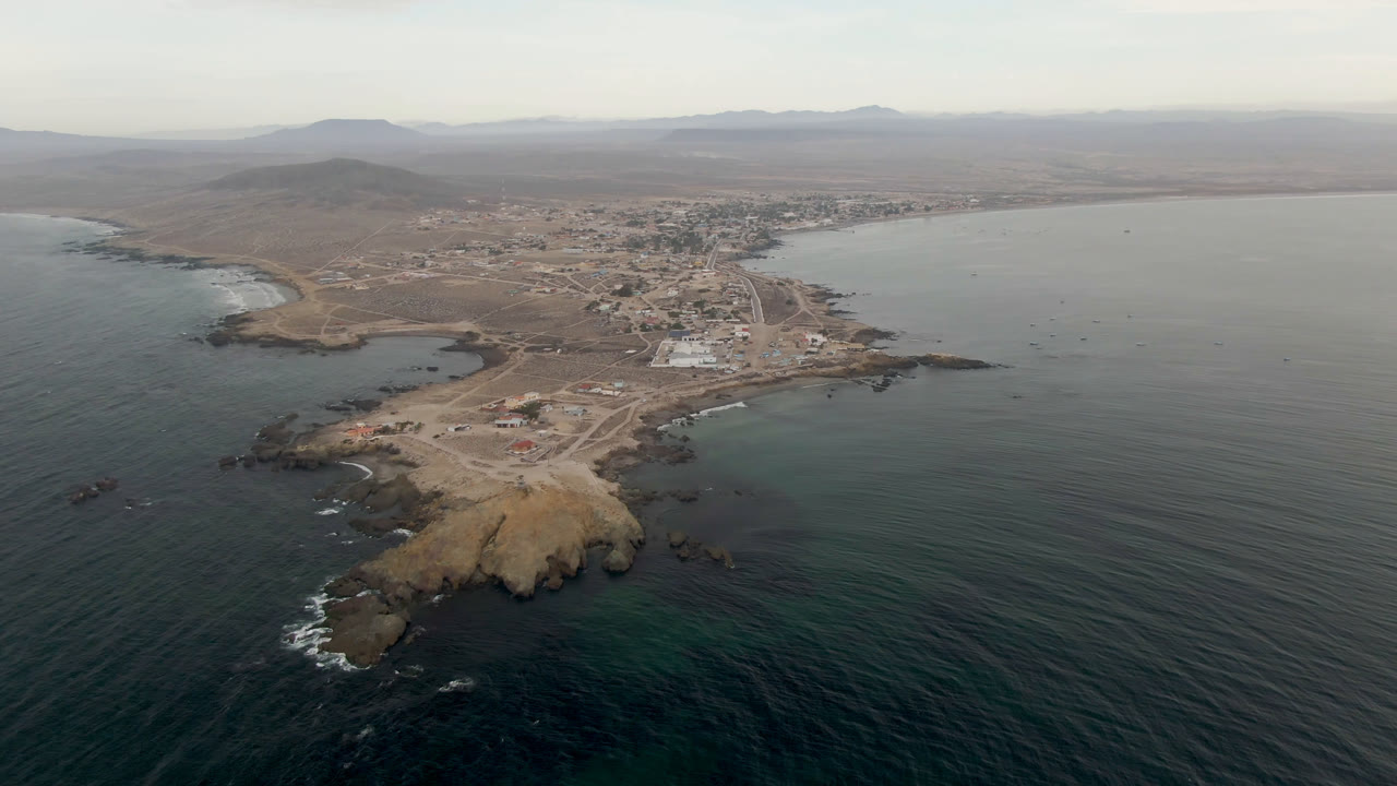 Aerial frame over Bahia Ascuncion, Baja California Sur, showing a rocky point in the foreground, dark ocean on both sides, a small town inland, hazy hills, and no visible people.