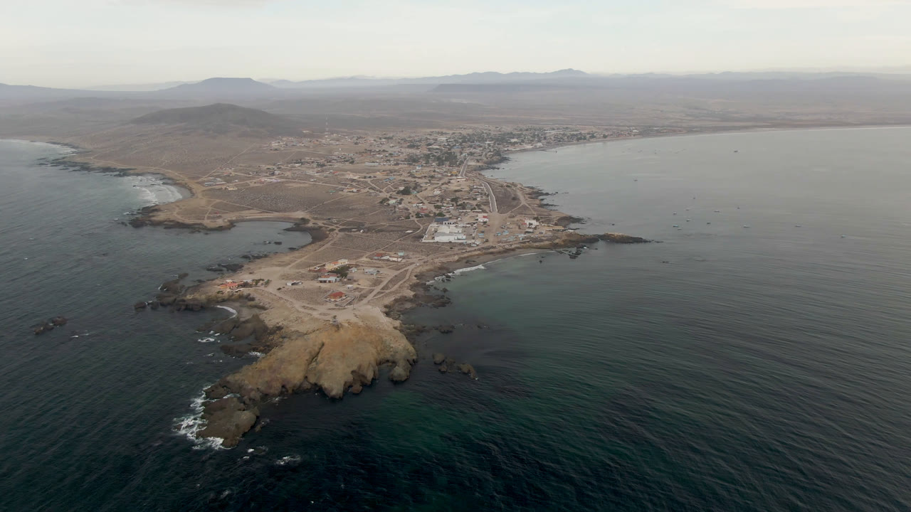 A high drone view of Bahia Ascuncion shows the rocky point slightly closer to center, with teal water in front, a curved bay to the right, town buildings behind it, and no visible people.