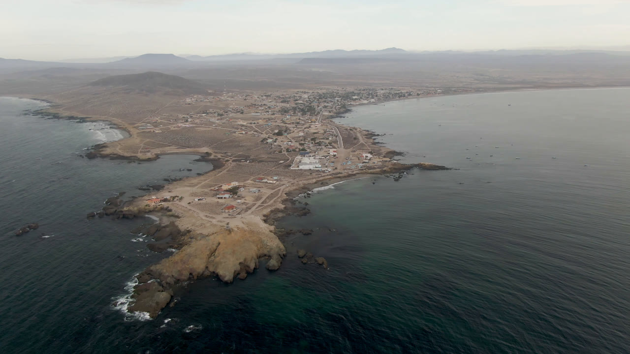 Aerial coastline with ocean, rocky shore, and town under hazy daylight
