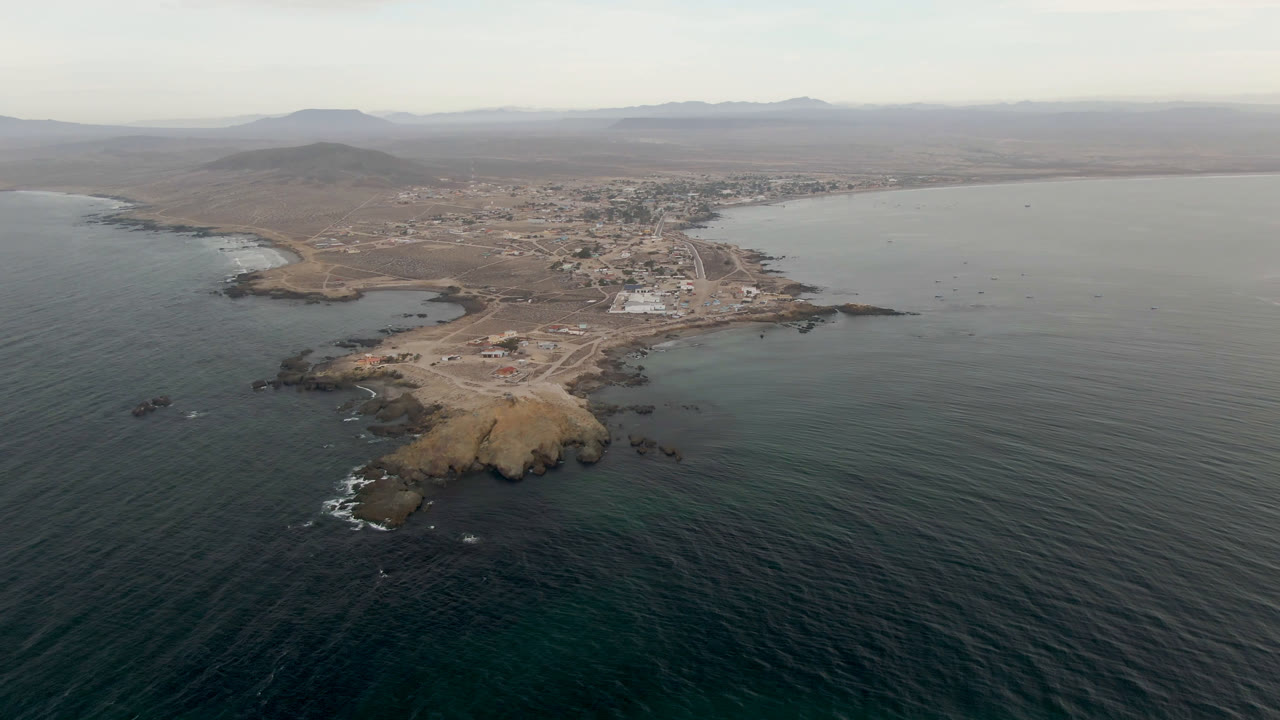 Aerial view of Bahia Ascuncion with the rocky shore slightly more centered, rippled water in front, low buildings inland, and no visible people.