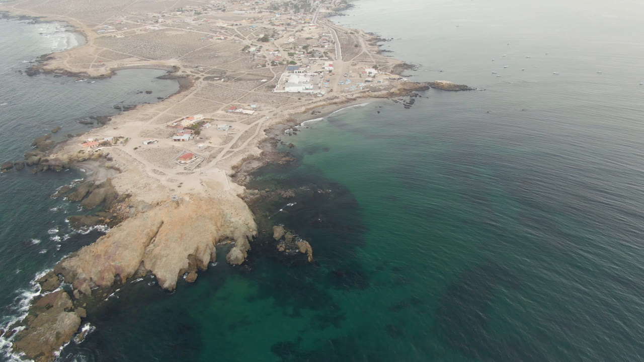 Aerial view in Bahia Ascuncion with the rocky point centered lower in frame, clear green-blue shallows on both sides, and a small town area visible near the upper edge, no visible people.