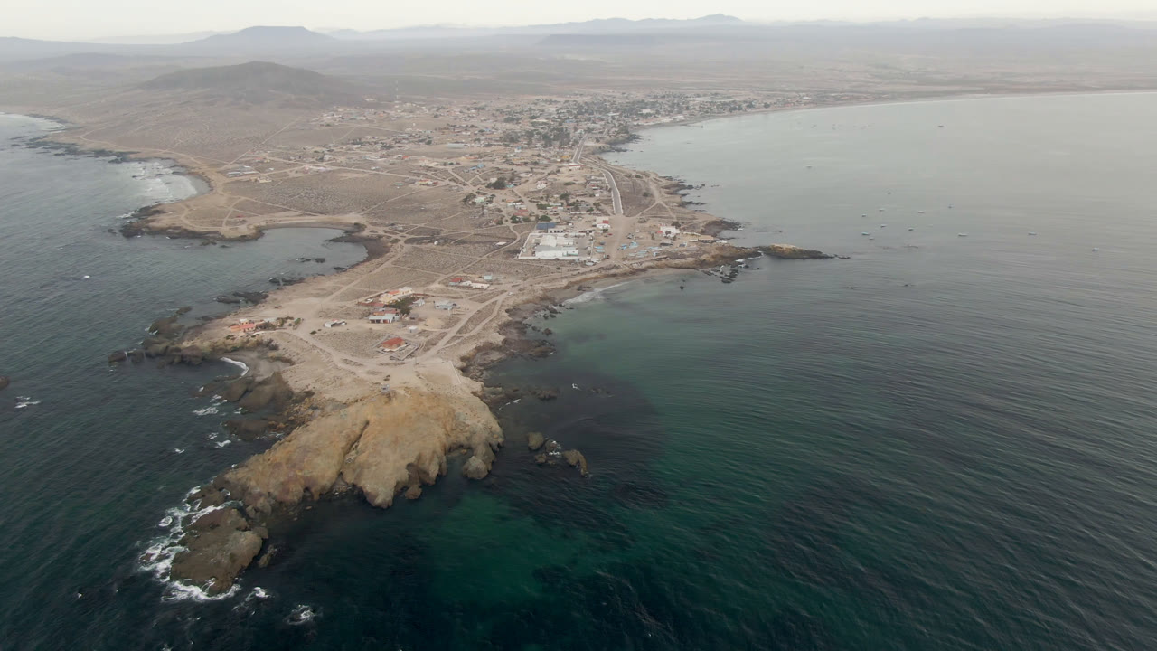 Aerial frame over Bahia Ascuncion showing a rocky point in the foreground, dark ocean and sheltered bay on both sides, low town buildings inland, and no visible people.