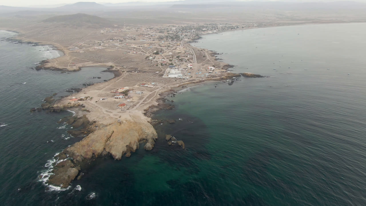 Aerial view in Bahia Ascuncion with the rocky point lower in frame, pale dirt roads and scattered buildings behind it, calm bay water to the right, and no visible people.