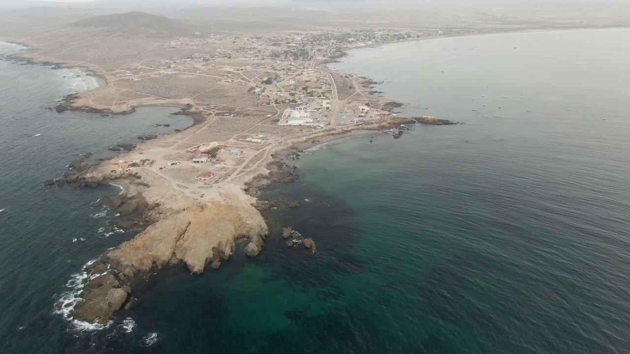 High aerial over dark blue ocean, rocky point, and small coastal town.