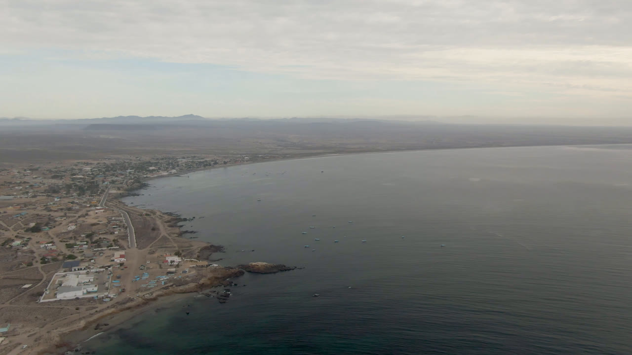Opening aerial frame over Bahia Ascuncion, Mexico, showing a curved shoreline, scattered buildings, dirt roads, calm water, distant hills, and no visible people.