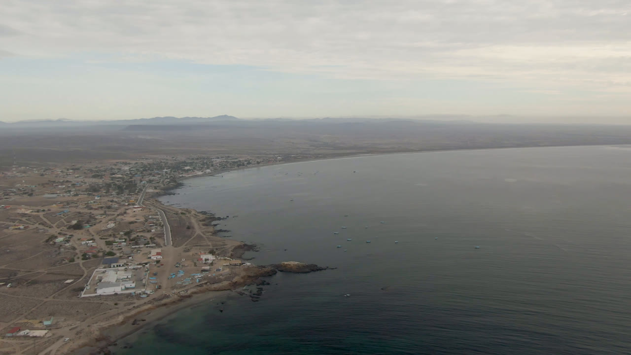 Aerial frame over Bahia Ascuncion, Baja California Sur, with a curving shoreline town at left, open bay water to the right, distant mountains, cloudy sky, and no visible people.