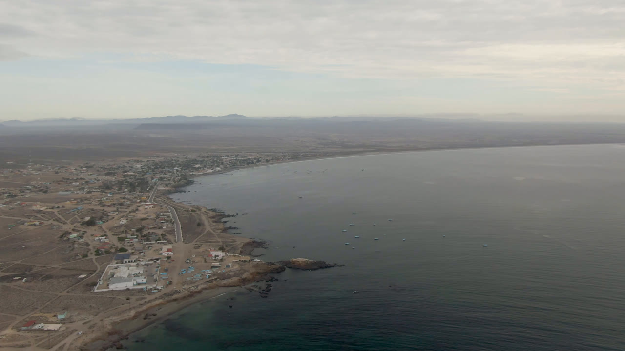 Second aerial frame of Bahia Ascuncion showing shoreline roads and low buildings along the left side, dark green-blue water across the right side, a low horizon, and no visible people.