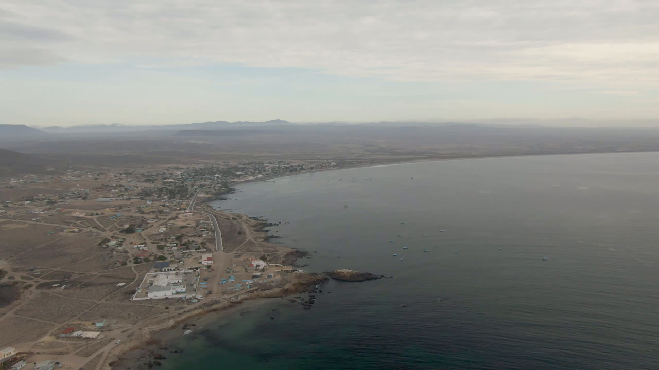 Aerial view over Bahia Ascuncion, Baja California Sur, Mexico, with roads and low buildings along the shore, dark water to the right, distant landforms on the horizon, and no visible people.