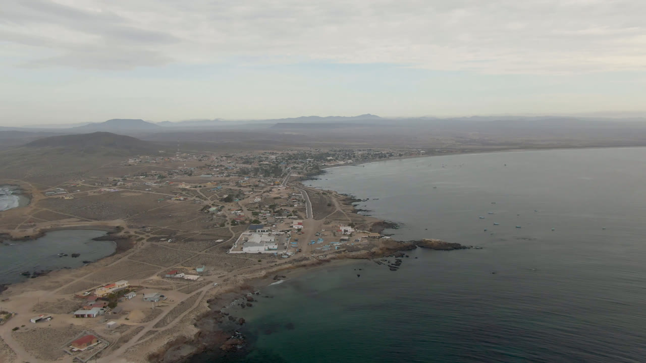 Aerial view in Bahia Ascuncion with a road leading toward scattered town buildings, rocky shore in the lower frame, calm bay water to the right, and no visible people.