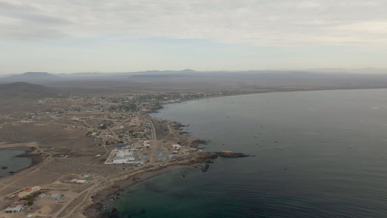 Aerial view of Bahia Ascuncion with the town closer to center, a curving shoreline, calm open water, low mountains on the horizon, cloud cover, and no visible people.