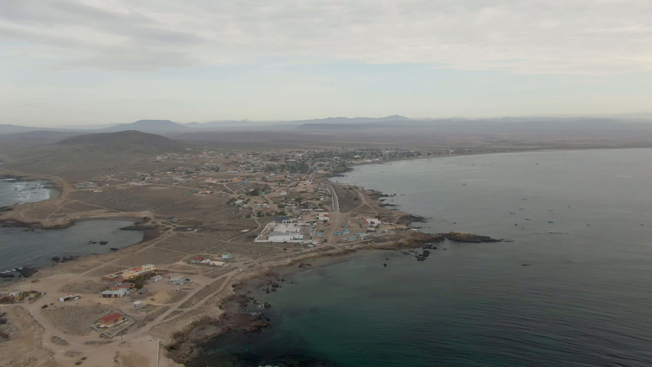 Aerial frame over Bahia Ascuncion, Baja California Sur, Mexico, showing dirt roads, scattered buildings, a rocky beach edge, broad bay water, distant hills, and no visible people at the start of the clip.