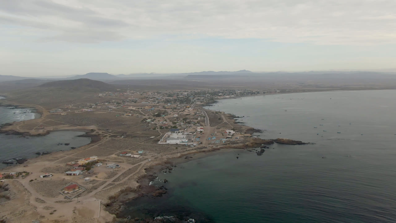 Wide aerial of bay water beside a small coastal settlement and inland hills.