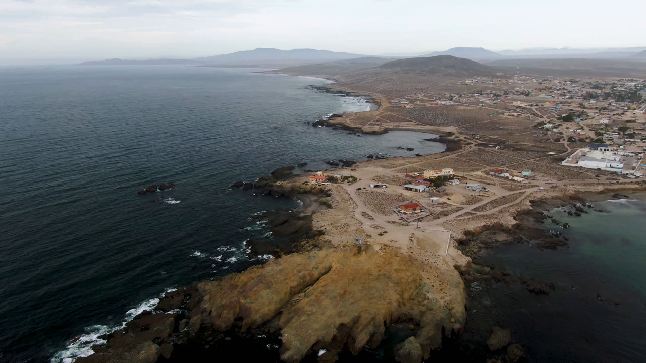 Aerial view of ocean, rocky point, dirt roads, and coastal town buildings.