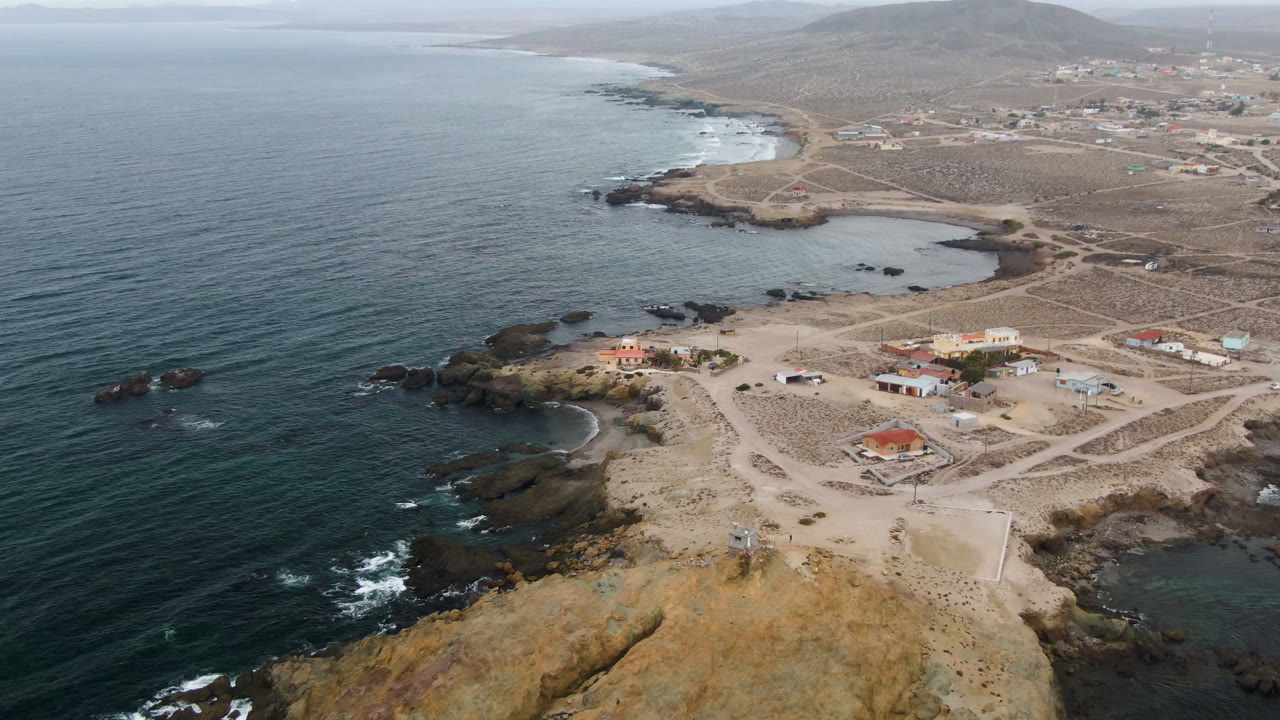 Aerial view over Bahia Ascuncion, Baja California Sur, showing dark ocean water beside a rocky point, pale sand roads, scattered low buildings, and no visible people.