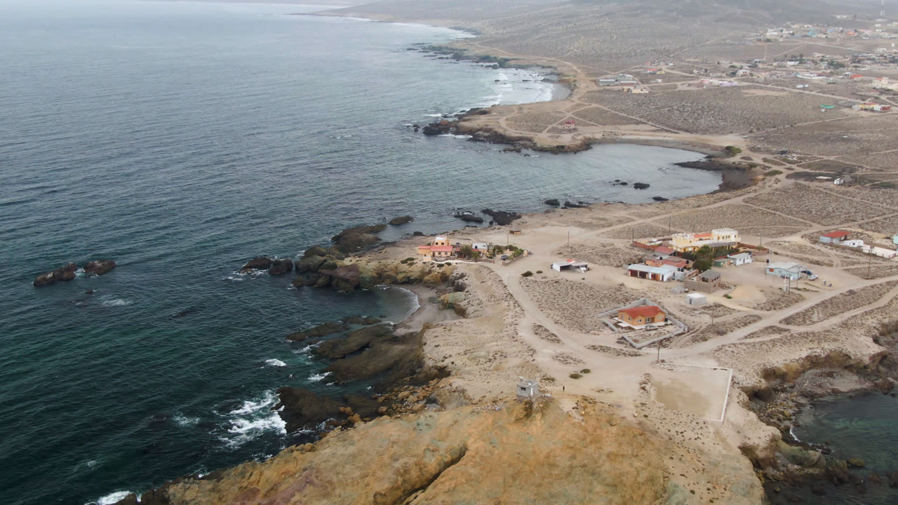Drone view over Bahia Ascuncion with the ocean on the left, a small cove and rock shelves below, sandy tracks through the settlement, and no visible people.