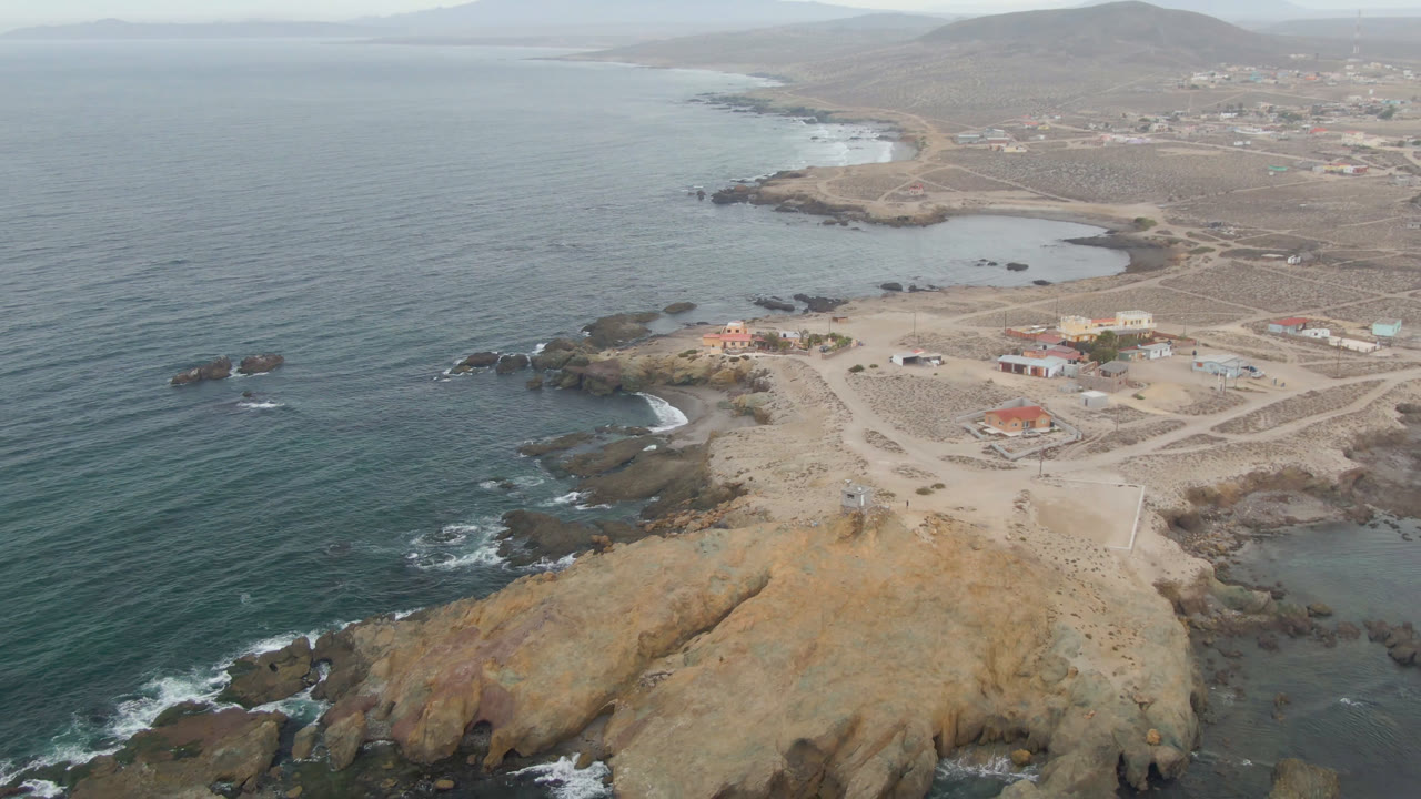 Drone view of rocky headland, ocean, sandy roads, and low coastal buildings