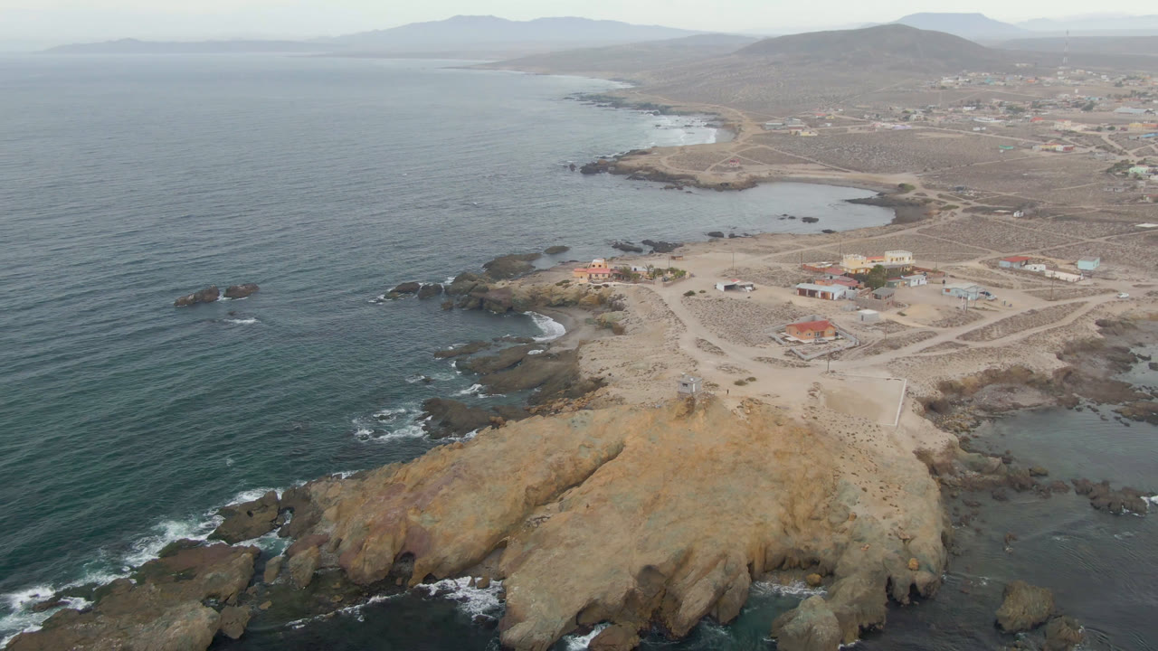 Drone frame over Bahia Ascuncion with the ocean occupying the left half, surf around offshore rocks, a tan point below, scattered buildings and roads inland, and no visible people.